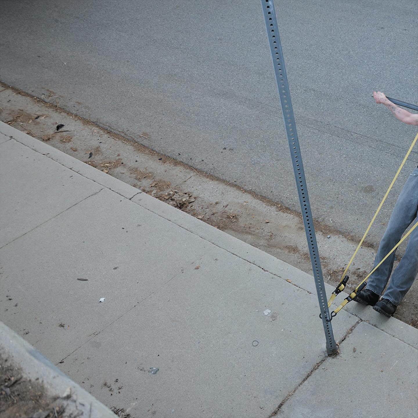 Corbin — Crisis Kid, street diptych (1 of 2): sign pole, the figure's legs and straps at the right edge