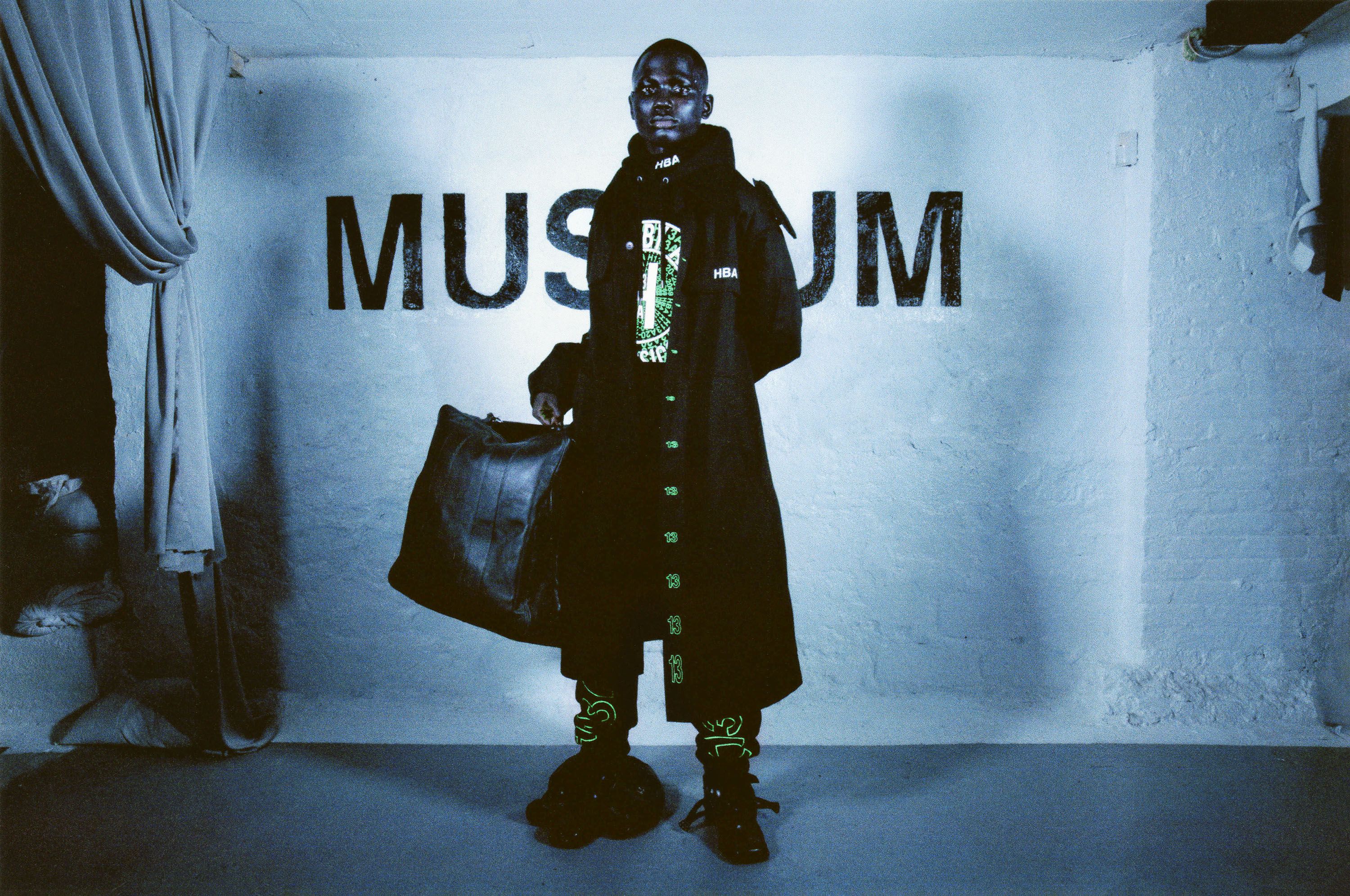 Hood By Air — Museum, model in a long dark coat before the Museum wall